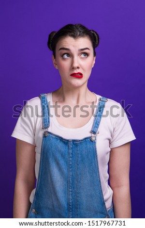 Portrait of woman doubting and confused, thinking of an idea or worried about something at studio. Girl with two hair-buns, wearing denim overalls and pink t-shirt.