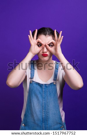 Pretty model looking at camera through her fingers in ok gesture. Imitating binoculars and beautiful eyes isolated over purple background at studio. Girl with two hair-buns