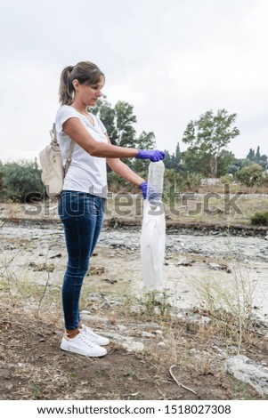 Stock photo of a girl standing with white t-shirt and jeans with blue gloves putting a plastic bottle in a bag. Action to recycle and clean nature