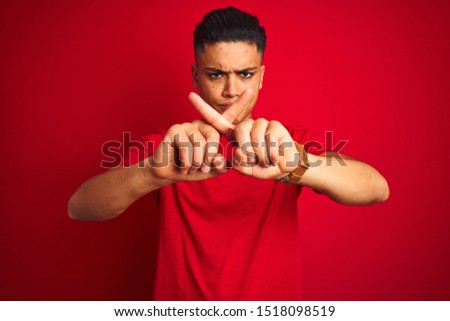 Young brazilian man wearing t-shirt standing over isolated red background Rejection expression crossing fingers doing negative sign