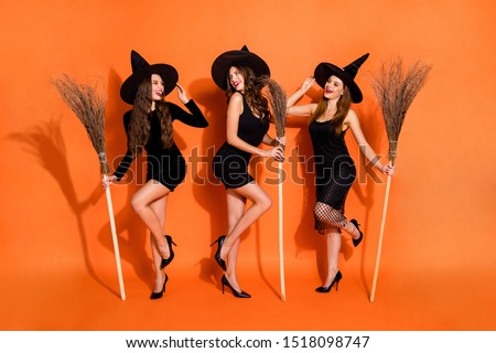 Full length photo of three witch ladies holding brooms talking about handsome wizards wear black dresses and hats isolated orange color background
