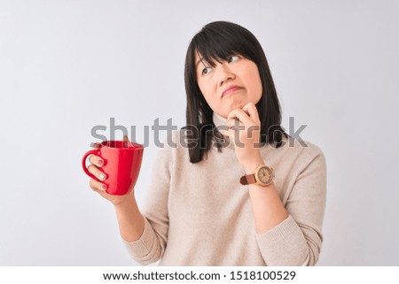 Young beautiful Chinese woman drinking red cup of coffee over isolated white background serious face thinking about question, very confused idea