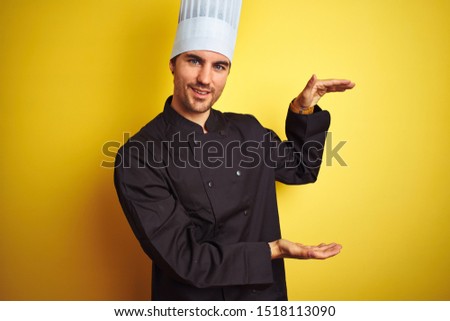 Young chef man wearing uniform and hat standing over isolated yellow background gesturing with hands showing big and large size sign, measure symbol. Smiling looking at the camera. Measuring concept.