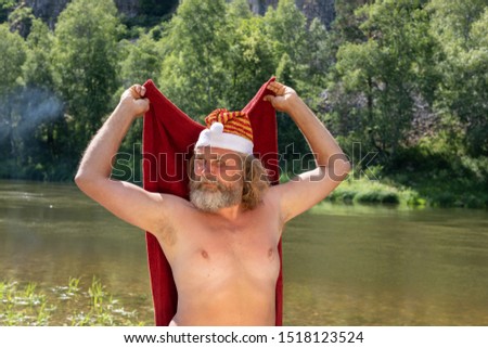 charismatic elderly man with a beard in a Santa Claus hat 
with a red blanket in his hands. Against the background of a summer landscape, river, mountains, forest