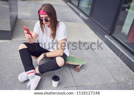 Photo of young brunette in sunglasses with phone in her hands sitting on skateboard on background of modern buildings in city