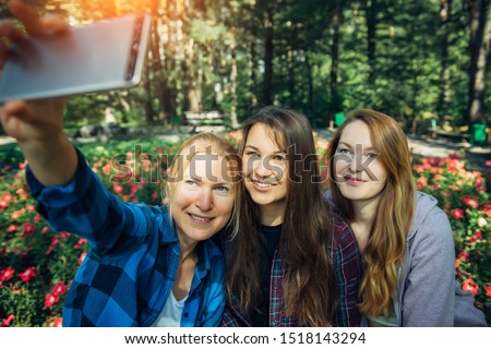 Three young cute girls taking selfie on smartphone in summer park. Happy women smiling at camera on flowering background. Friendship concept.