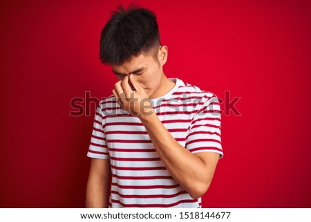 Young asian chinese man wearing striped t-shirt standing over isolated red background tired rubbing nose and eyes feeling fatigue and headache. Stress and frustration concept.