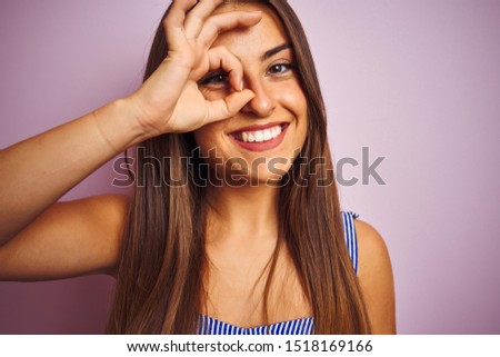 Young beautiful woman wearing t-shirt standing over isolated pink background with happy face smiling doing ok sign with hand on eye looking through fingers