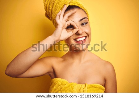 Young beautiful woman wearing towel after shower over isolated yellow background with happy face smiling doing ok sign with hand on eye looking through fingers