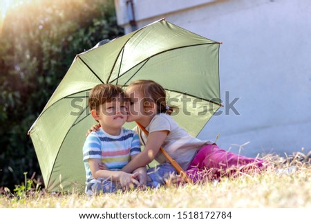 Sibling love.Beautiful blond girl is kissing little brother under umbrella.Two fun,happy children playing and enjoying the sunny day in the garden.Brother and sister under green umbrella in the park.
