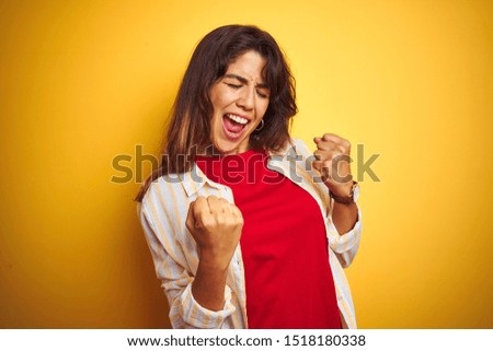 Young beautiful woman wearing red t-shirt and stripes shirt over yellow isolated background very happy and excited doing winner gesture with arms raised, smiling and screaming for success. 