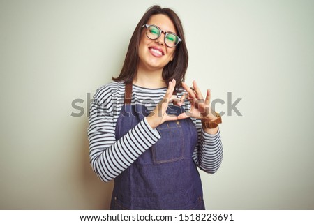 Young beautiful business woman wearing store uniform apron over isolated background smiling in love showing heart symbol and shape with hands. Romantic concept.