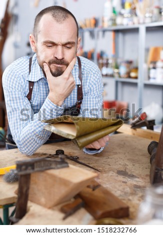 Annoyed workman dissatisfied with quality of leather for upholstering chair in repair furniture workshop