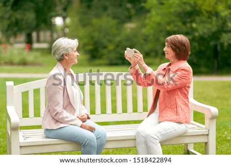 old age, retirement and people concept - senior woman photographing her friend sitting on bench at summer park by smartphone