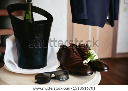 Close up of brown male shoes, boutonniere, sunglasses and champagne in bucket on white wood table, copy space. Modern men accessories. Wedding details 