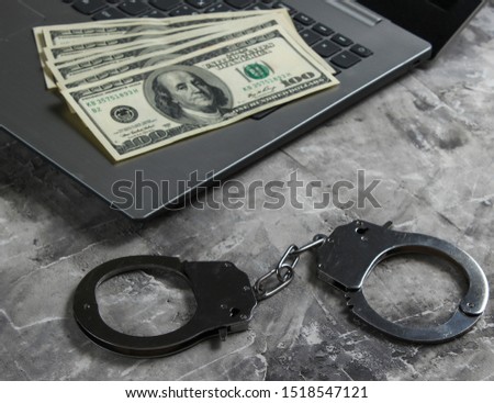 Cybercrime, online digital theft. Laptop with dollar bills and
 steel handcuffs on a gray concrete background. 