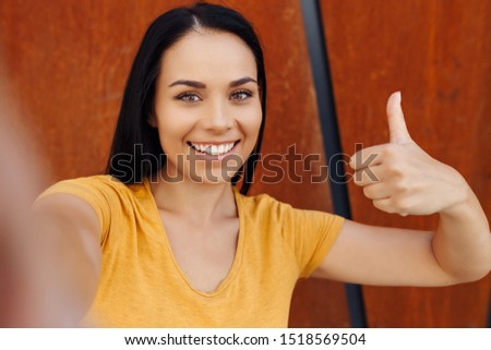 Funny brunette young woman with bright smile dressed in casual clothes doing selfie on smartphone and show ok gesture in front of rusty wall.