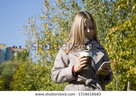 A young pretty woman looking at her phone and holding a cup of coffee in park