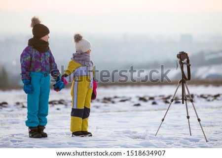 Two children boy and girl having fun outside in winter playing with photo camera on a tripod on snow covered field.