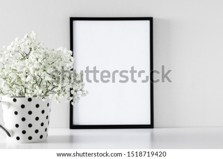 Black frame photo on white wood table and small white flowers in vase. White gypsophila flowers on shelf or desk. Mock up with decor elements.