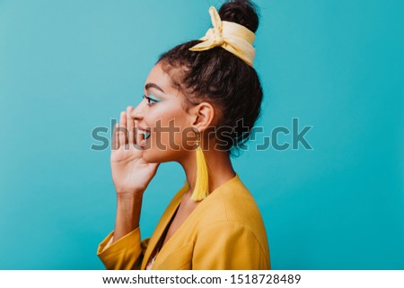 Attractive brunette woman with long yellow earrings posing in studio. Indoor photo of good-humoured african lady isolated on blue background.