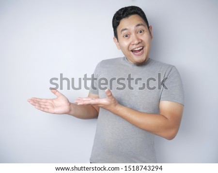Portrait of funny young Asian man smiling and pointing to presenting something on his side, against grey background with copy space