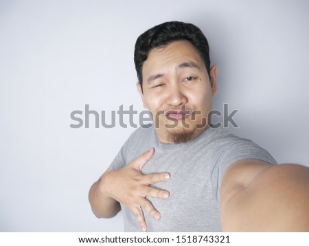 Portrait of young Asian man smiling while taking selfie photograph of himself on his smart phone with funny expression