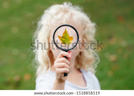 Little child girl holding magnifying glass and covering her face. Wet autumn fall maple leaf on glass with rain water drops. Kid playing outdoor. Happy childhood, education and ecology concept.