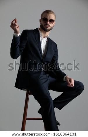 attractive formal business man wearing a navy suit and sunglasses sitting with one leg resting on a chair and one hand raised with two fingers