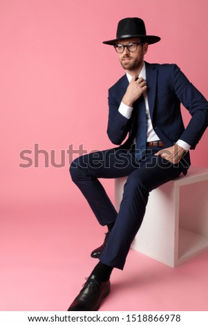 mysterious formal business man wearing a navy suit,black hat,glasses is sitting and resting his hand on his lap while fixing his tie and looking away happy on pink studio background
