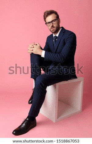 good looking formal business man wearing a navy suit and  glasses sitting and holding one leg resting on a chair and looking at camera serious on pink studio background