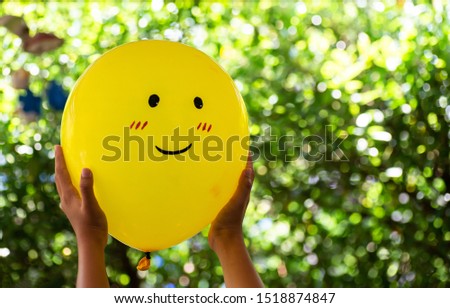 Hands holding smiling yellow balloon on dark green hedge leaves background outdoors. Happy emotions concept. 