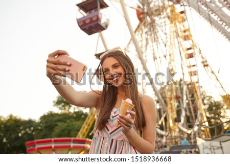 Young beautiful young woman with brown hair posing over ferris wheel on sunny warm day, wearing sunglasses and romantic dress, smiling cheerfully to camera while making selfie with smartphone