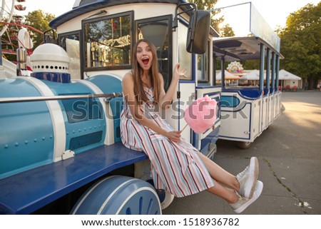 Outdoor shot of cheerful attractive lady in light long dress sitting on steam train in amusement park on warm summer day, posing to camera with wide mouth opened and pink cotton candy in hand