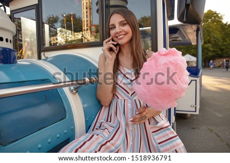 Young lovely long haired female in romantic dress sitting in amusement park and leaning on fake steam train, having pleasant talk on phone and smiling cheerfully to camera with cotton candy in hand