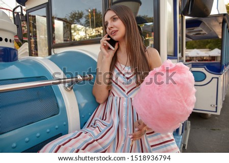 Beautiful young brunette in light summer dress sitting over amusement park with cotton candy on wooden stick, making call with her smartphone, waiting for her friends