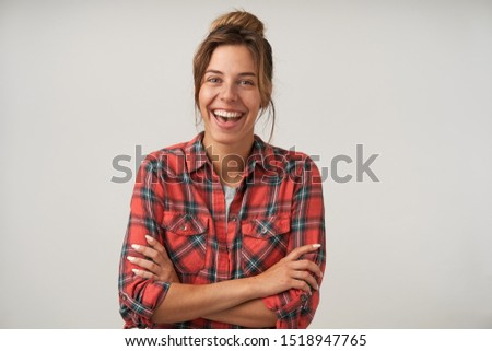 Happy young attractive female with bun hairstyle posing over white background, smiling joyfully to camera with crossed hands on her chest, positive emotions concept