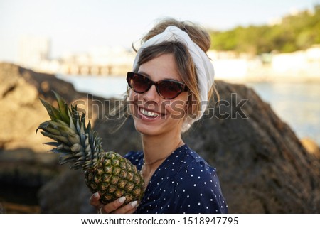 Beautiful young dark haired woman with casual hairstyle wearing headband and looking at camera with broad happy smile, posing over sea bay on sunny summer day with fresh pineapple in hand