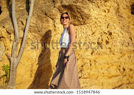 Outdoor shot of pretty dark haired woman in casual clothes standing over yellow stone wall, looking at camera with happy smily and holding her skirt with hands