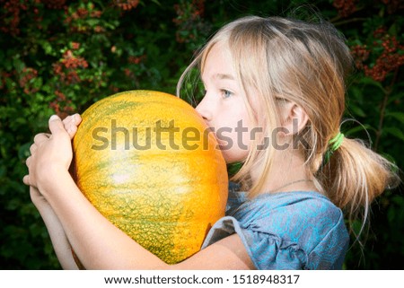 Cute blond child girl holding and kissing pumpkin outside in garden with greenery background