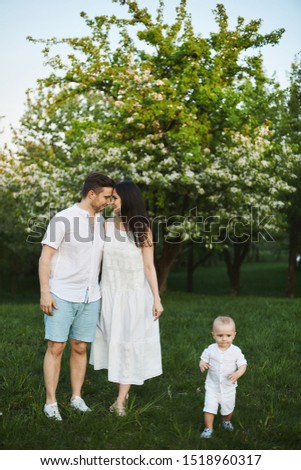 Beautiful young parents, stylish handsome man in shorts and shirt and brunette beauty in the white dress, and their cute little son have fun near trees at blooming park. Concept of a happy family
