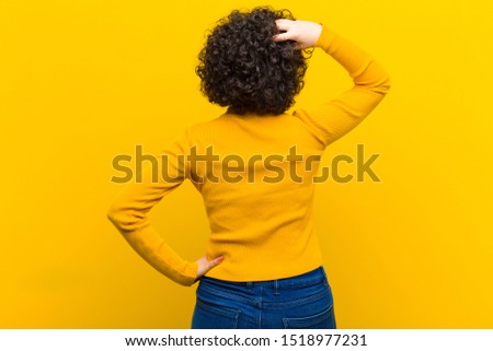 young pretty afro woman feeling clueless and confused, thinking a solution, with hand on hip and other on head, rear view