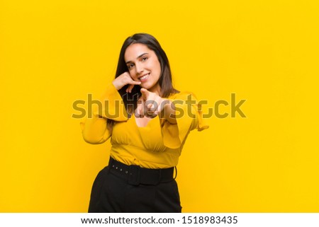 young pretty woman smiling cheerfully and pointing to camera while making a call you later gesture, talking on phone isolated against  wall