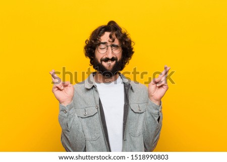 young crazy man smiling and anxiously crossing both fingers, feeling worried and wishing or hoping for good luck against yellow wall