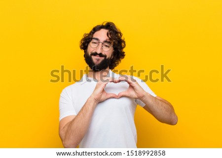 young crazy man smiling and feeling happy, cute, romantic and in love, making heart shape with both hands against yellow wall