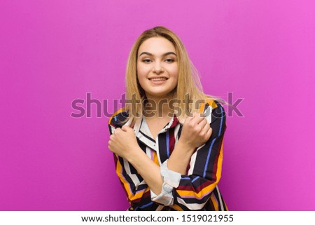 young blonde woman smiling cheerfully and celebrating, with fists clenched and arms crossed, feeling happy and positive against purple wall