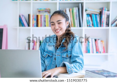 Picture of a female teenager smiling at the camera while studying with a computer laptop in the library with bookcase background