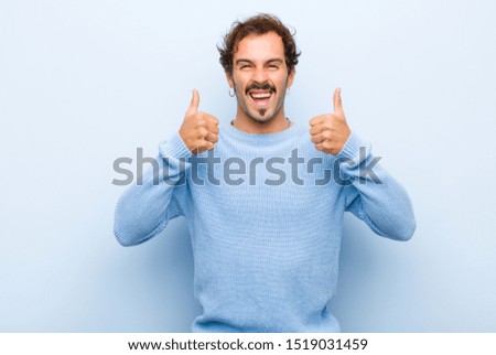 young handsome man smiling broadly looking happy, positive, confident and successful, with both thumbs up isolated against flat wall