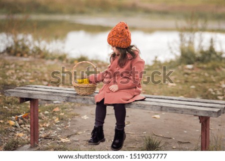 sad little preschooler girl in a brown coat and orange hat sits on a wooden bench near the lake, looks at the basket to the left. horizontal photograph of a child.