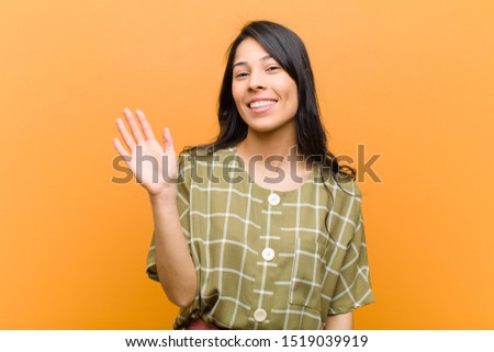 young pretty hispanic woman smiling happily and cheerfully, waving hand, welcoming and greeting you, or saying goodbye against brown wall
